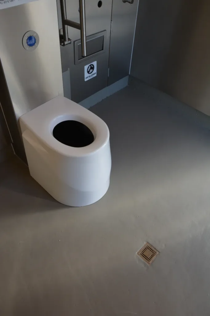 Nahaufnahme des Bodens in einer Kubus-Komforttoilette mit Trocken-Trenntoilette ECODOMEO: glatter, grauer Betonboden, der bauseitig vom Kunden hergestellt wird, weißes Toilettenbecken, Edelstahl-Haltegriff, saubere und ebene Ausführung
