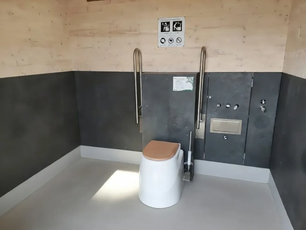 Innenraum einer barrierefreien Kubus-Komforttoilette mit ECODOMEO Trocken-Trenntoilette (wasserlos mit Förderband): weißer Toilettensitz mit Trenneinsatz, zwei Edelstahl-Haltegriffe für barrierefreien Zugang, untere Wandverkleidung aus grauen Hanit-Platten, obere Verkleidung aus heller Dreischichtplatte. Infotafel mit Piktogrammen zu Benutzung und Hygiene, heller Holzboden