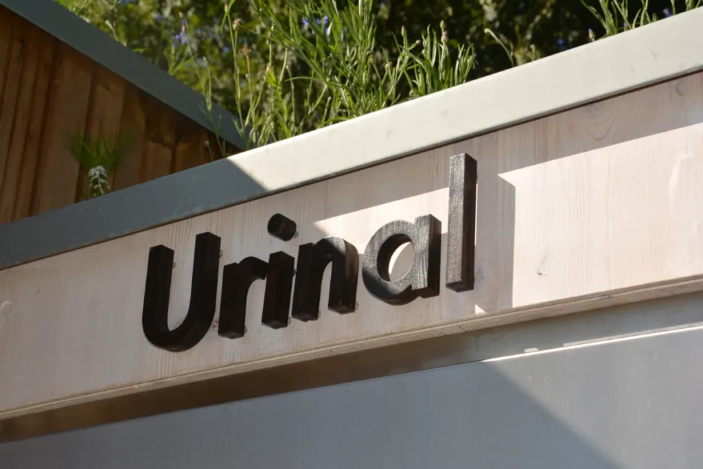 Beschilderung „Urinal“ an der öffentlichen Toilettenanlage KUBUS: klare Beschriftung für separaten Urinalraum, Gründach mit Pflanzen für nachhaltige Optik und Kühlung, Lärchenholz-Design, barrierefrei nach DIN 18040, umweltfreundlich und einladend