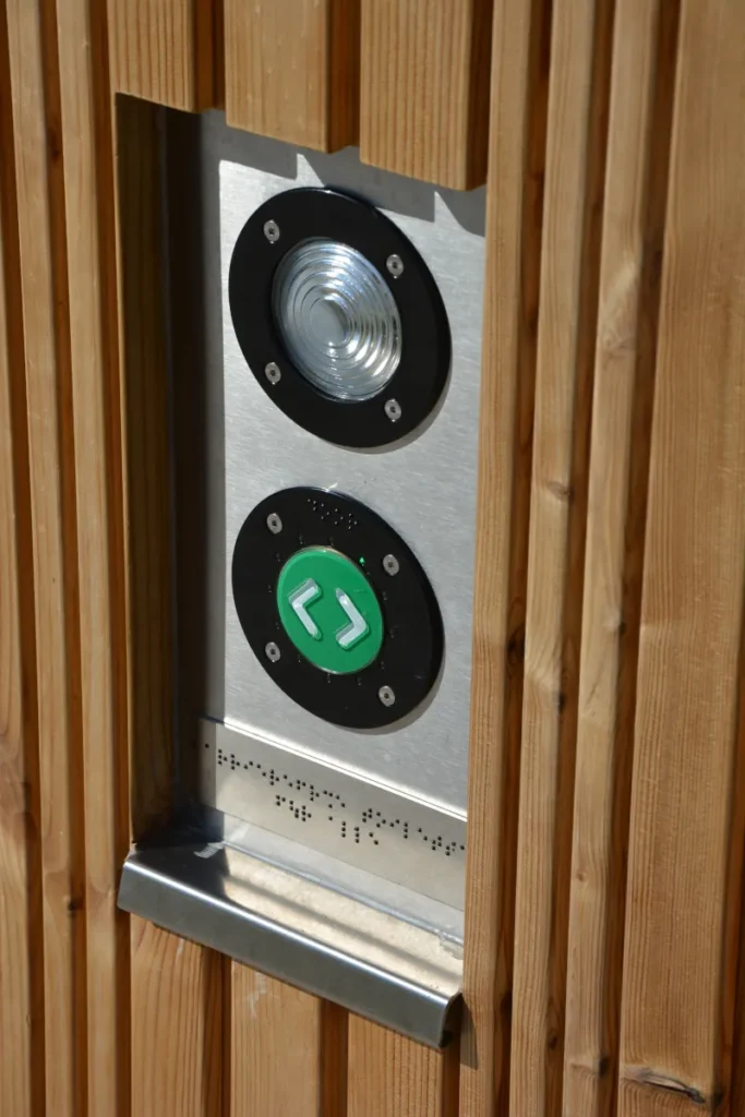 Toilettenanlage KUBUS - Bedienelement für elektrische Türöffnung per Knopfdruck in barrierefreier Toilettenanlage KUBUS: taktile Brailleschrift-Beschriftung für blinde und sehbehinderte Personen, gut ertastbar, inklusiv und DIN 18040-konform, kombiniert mit visueller Beschriftung für maximale Zugänglichkeit