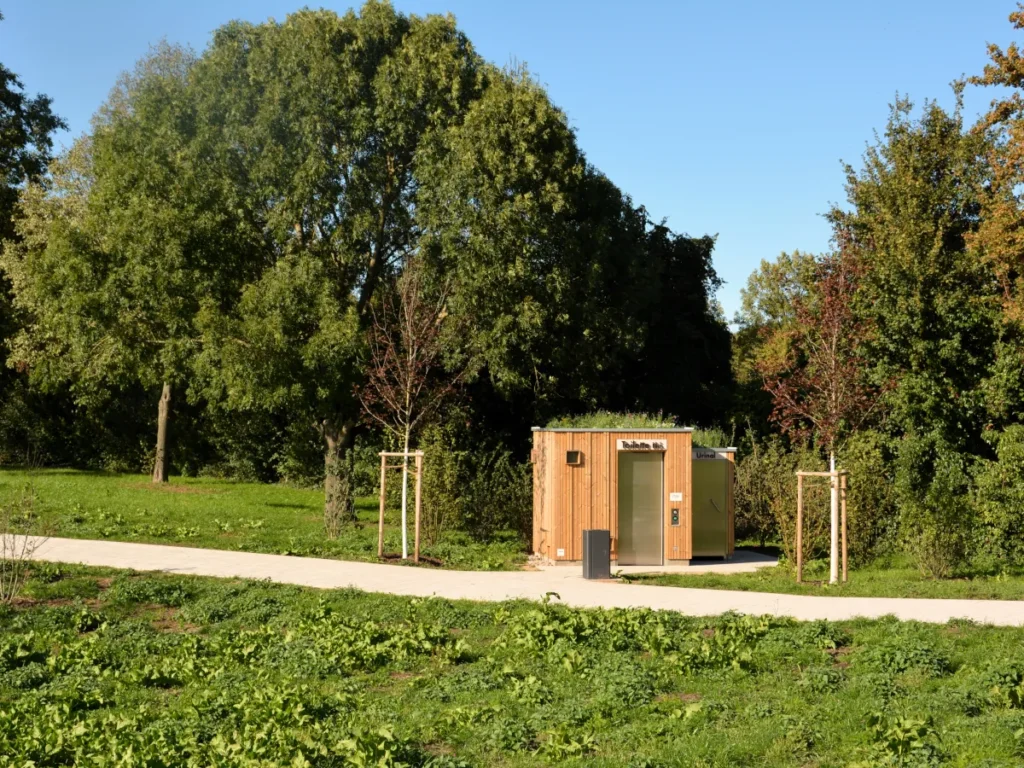 orderansicht der barrierefreien Toilettenanlage KUBUS nach DIN 18040 in einem Park: Lärchenholz-Fassade, Eingang mit Schiebetür, optionales Gründach, vollständig autark ohne Wasser- oder Kanalisationsanschluss – nachhaltig, einladend und perfekt in Grünanlagen integriert, ideal für öffentliche Plätze, Wanderwege und Rastplätze