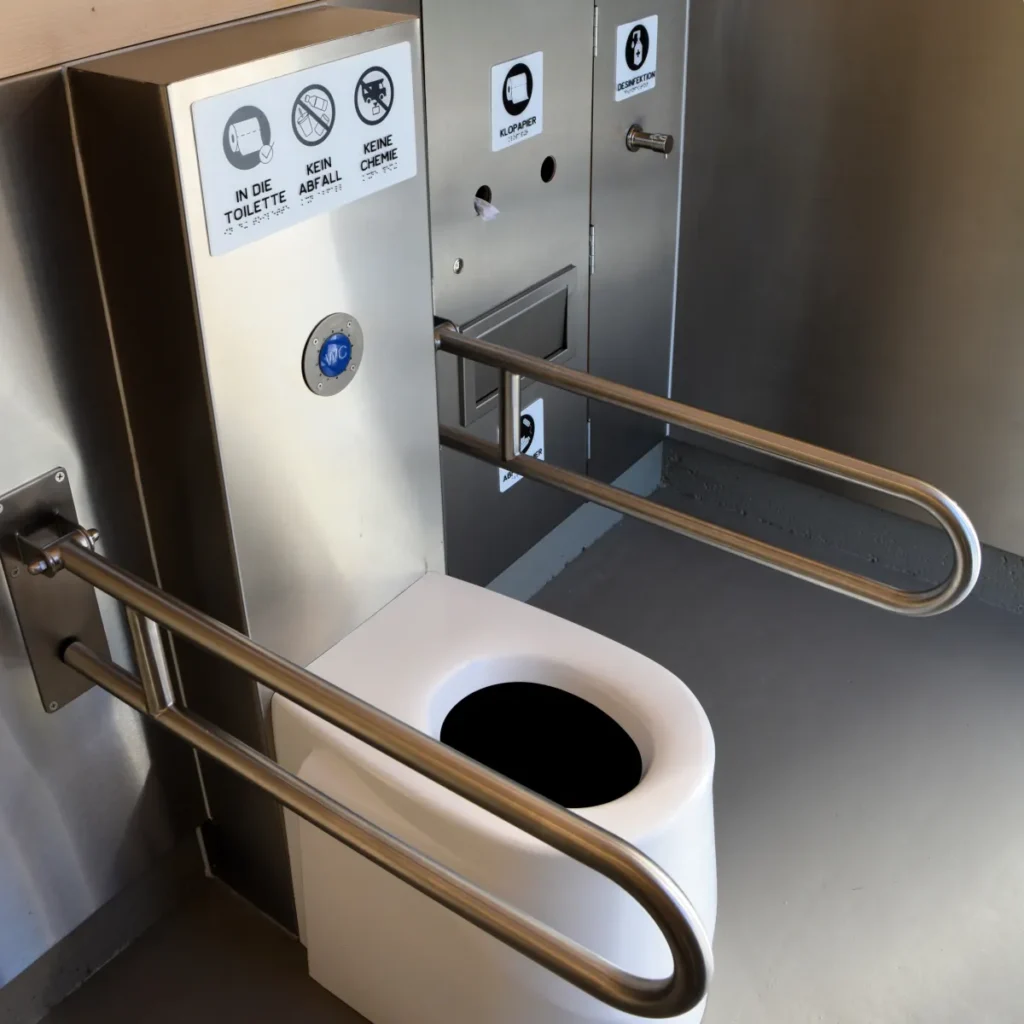Detailansicht des Innenraums einer öffentlichen, barrierefreien Toilette (nach DIN 18040): ECODOMEO Trockentrenntoilettensystem mit automatischem Förderband für geruchlose Trennung von Urin und Fäkalien, integrierte wandbündige Spender (Toilettenpapier, Handgel, Mülleimer) vandalensicher, klappbare Edelstahl-Stützgriffe für barrierefreie Nutzung