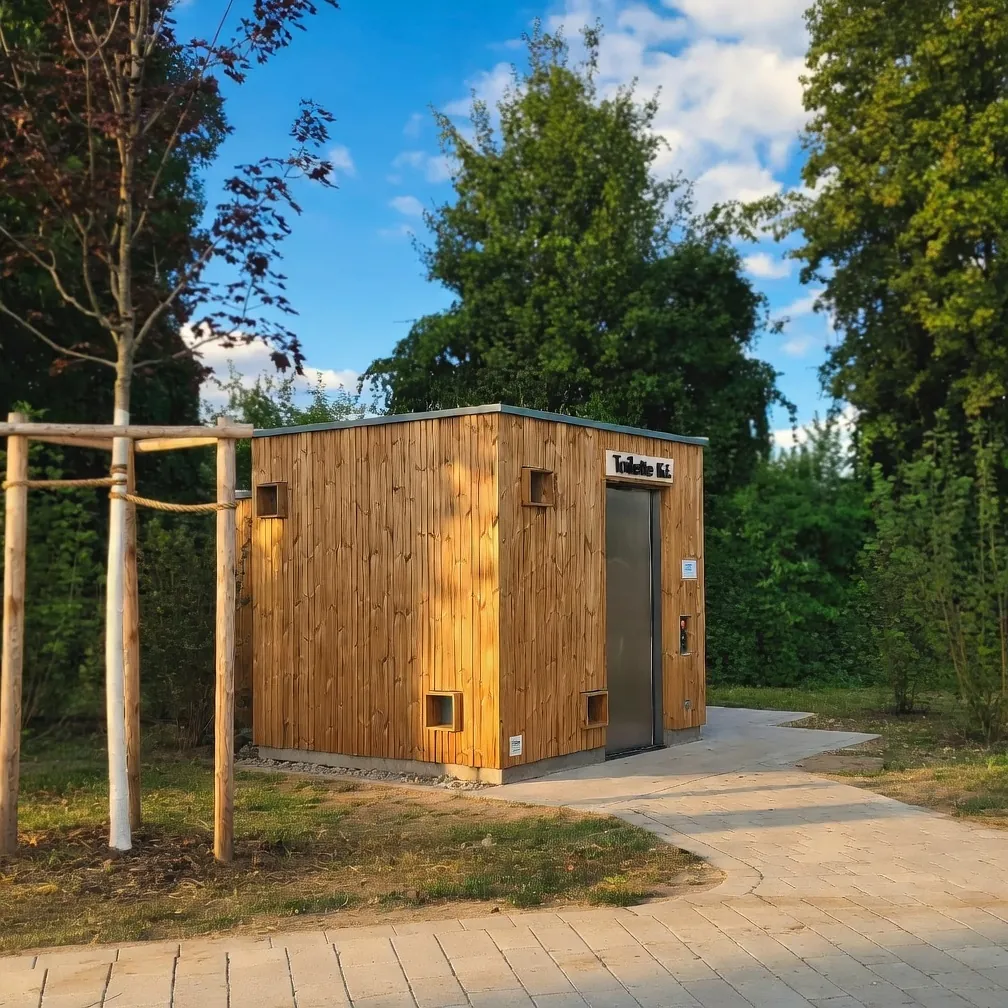 Moderne Holzkabine KUBUS als öffentliche Trockentoilette in einer Parkanlage in Hanau, natürliches Holzdesign, Edelstahltür, kleine Lüftungsöffnungen und strahlend blauer Frühlingshimmel im Hintergrund.