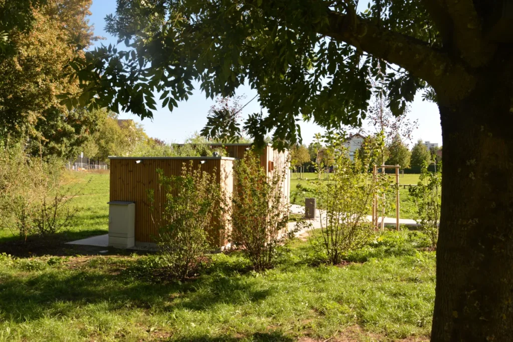 Hinteransicht der öffentlichen KUBUS-Toilettenanlage in parkähnlicher Umgebung: massive Lärchenholz-Verkleidung, optionales Gründach für Biodiversität und Kühlung, vollständig unabhängig von Wasser- oder Abwasseranschlüssen (Erdtank + Solar), barrierefrei nach DIN 18040, umweltfreundlich und flexibel einsetzbar ohne Infrastruktur